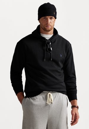 Zwarte hoodie met een klein logo, gedragen met grijze joggingbroeken. Accessoires omvatten een zwarte gebreide beanie en een zonnebril die om de nek hangt.