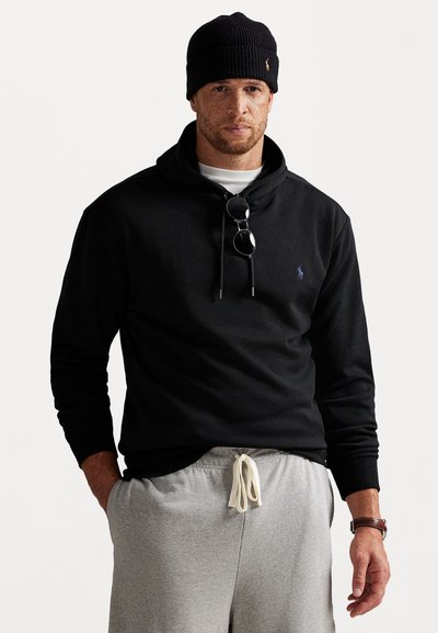 Sudadera negra con capucha y un pequeño logo, combinada con pantalones de chándal grises. Los accesorios incluyen un gorro de lana negro y unas gafas de sol colgadas del cuello.