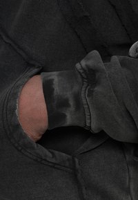 Μαύρο φούτερ με φθαρμένη σχεδίαση, ορατή ραφή και χέρι εντός στενού πλεκτού μανσετιού, παρουσιάζοντας ποικίλες υφές.