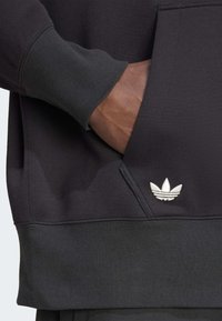 Sudadera negra de tejido suave, con un bolsillo tipo canguro, puños acanalados y un logotipo de Adidas bordado en blanco en el bolsillo.