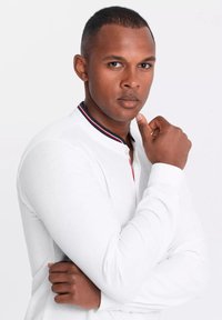 Camisa blanca de manga larga con cuello, que presenta un acento en rojo y azul marino en el cuello y una tela texturizada. El modelo posa con los brazos cruzados.