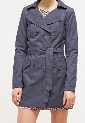 Mujer que lleva un abrigo trench azul marino de doble botonadura con un cinturón atado y un vestido floral que asoma por debajo.