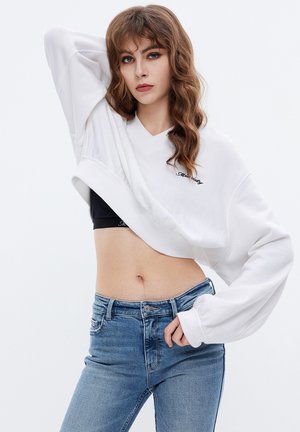 Hvid cropped sweatshirt med v-hals-design, ribbede manchetter og broderet tekst. Stylet med blå højtaljede jeans, der fremhæver en afslappet stil.