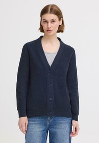Gilet bleu marine avec un motif de côtes texturé, col en V et six boutons assortis. Porté par-dessus un haut gris, manches longues visibles.
