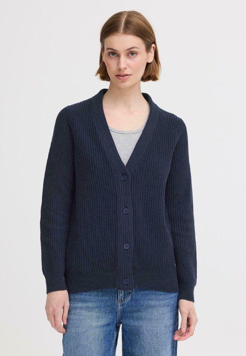 Gilet bleu marine avec un motif de côtes texturé, col en V et six boutons assortis. Porté par-dessus un haut gris, manches longues visibles.