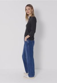 Svart långärmad tröja med vit krage, i kombination med vida blå denimjeans och vita sneakers. Modellen står sidleds mot en enkel bakgrund.