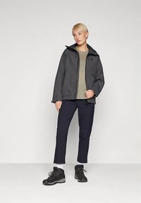 Black waterproof jacket with a zip, hood, and textured finish, worn over a green shirt, paired with dark pants and black hiking boots.