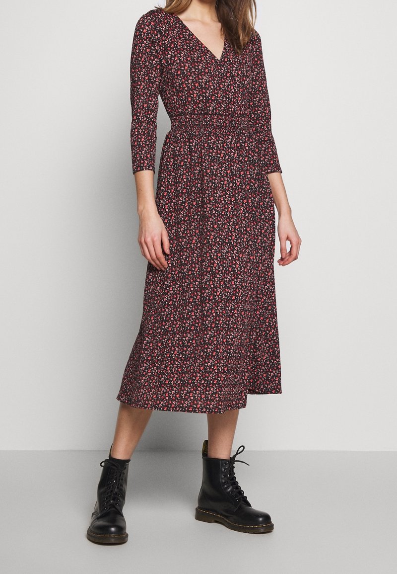 Frau in einem schwarz-roten Blumenkleid mit langen Ärmeln und tailliertem Schnitt, dazu schwarze Schnür-Ankle-Boots, steht auf einem grauen Boden.