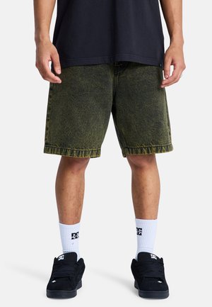 Man met een zwart t-shirt, vervaagde donkergroene knielange shorts, witte sokken met logo en zwarte sneakers, staand tegen een witte achtergrond.