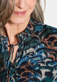 Donna che indossa una blusa a fantasia con colletto arricciato e dettaglio a fiocco, caratterizzata da un design astratto blu, verde, nero e marrone.
