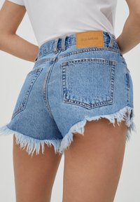 Shorts de denim con un lavado azul claro, dobladillo deshilachado y parche de cuero marrón de Pull&Bear en la cintura. Dos bolsillos traseros visibles.