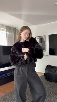 Czarna kurtka z czarnego futerka z długimi rękawami i krótkim fasonem, zestawiona z czarnymi spodniami ze ściągaczem. Gładka faktura, bez widocznych wzorów.