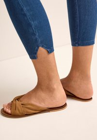 Sandalias marrones tipo slip-on con detalle de nudo, combinadas con pantalones de mezclilla azul de longitud hasta el tobillo que tienen un corte en el dobladillo.