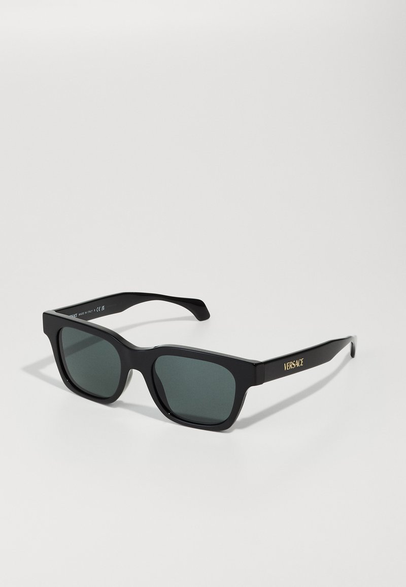 Gafas de sol de cuadrado negro con lentes grises, que presentan una textura de plástico suave y un logo dorado "VERSACE" en las patillas.