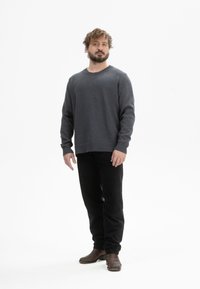 Mann mit Bart, der einen dunkelgrauen Pullover, schwarze Hosen und braune Stiefel trägt, steht frontal vor einem weißen Hintergrund.
