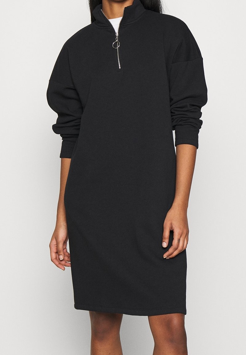 Femme portant une robe noire à manches longues, longueur genou, avec un col demi-fermeture éclair et un tire-zip en anneau, debout sur un fond uni.