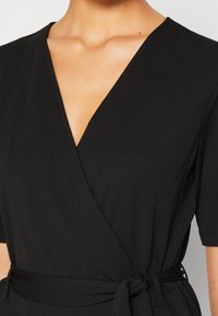 Mujer con un vestido negro cruzado de mangas cortas y un cinturón atado a la cintura, mostrando el escote y las clavículas.