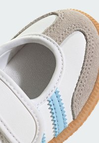 Biele a šedé tenisky s nubukovým zvrškom, modrými akcentmi a textúrovanou podrážkou. Zapínanie obsahuje široký suchý zips.