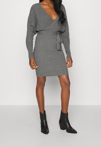 Robe enveloppante grise en tricot avec des manches longues, un décolleté en V et une ceinture à la taille. Bottines noires à bout pointu et à talon épais.