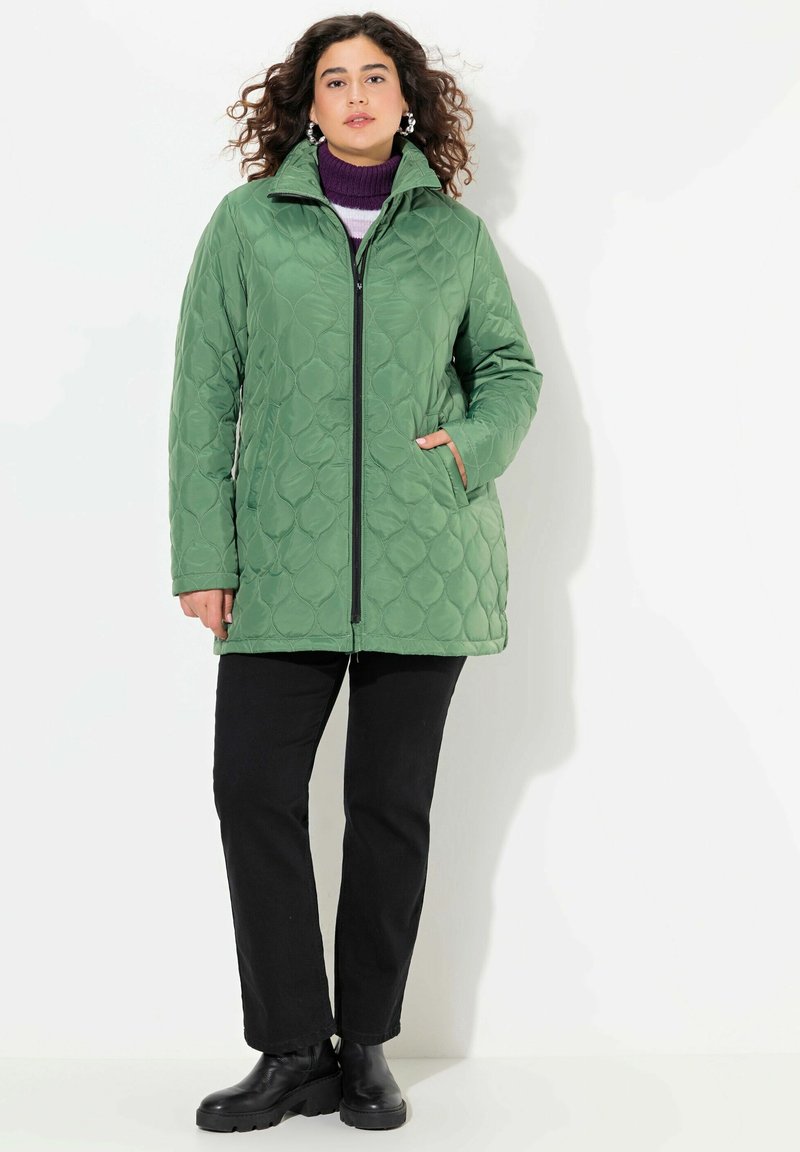 Prošívaná zelená bunda s vysokým límcem, zipovým zapínáním a bočními kapsami. Kombinováno s černými kalhotami a černými lodenými botami.