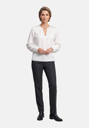 Donna in piedi che indossa una camicetta bianca a maniche lunghe, pantaloni dritti grigio scuro e stivaletti neri, su sfondo semplice.