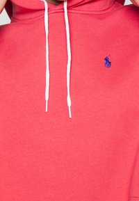 Korálová mikina vyrobená z měkké tkaniny s klokánkovou kapsou, s bílými stahovacími šňůrkami a modrým vyšitým logem na levém hrudi.