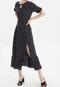 Robe maxi noire à pois avec des manches courtes bouffantes, un décolleté en trou de serrure, une fente sur le côté et un ourlet à volants. Modèle portant des sandales à talons ouverts noires.