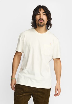 Homme aux cheveux foncés mi-longs et à la barbe, portant un t-shirt blanc avec une petite figure brodée sur la poche poitrine et un pantalon marron.