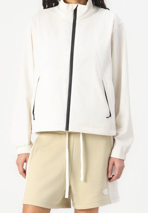 Person iført en hvid lynlåstrøje med sorte lynlåse og beige shorts med snøre mod en ensfarvet hvid baggrund.