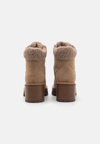 Botas de tobillo de ante marrón con un puño de piel de faux beige texturizado, tacón de madera grueso y suela lisa. La vista trasera muestra los detalles de la costura.