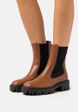 Bruna läderankelboots med elastiska sidopaneler, tjock svart gummisula och robust mönstring. Slät textur med minimala sömmar och detaljer.