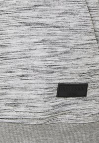 Light gray sweater with a textured, heathered pattern. Features a black logo patch labeled "BRAVESOUL LONDON" at the lower left.