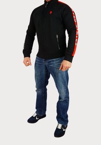 Giacca nera con zip e dettagli rossi, logo sul petto; abbinata a jeans in denim blu e sneakers nere con lacci bianchi.