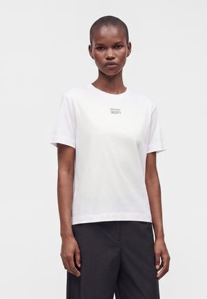 Femme aux cheveux courts portant un T-shirt blanc à manches courtes avec un petit texte sur la poitrine et un pantalon noir, debout devant un fond blanc.