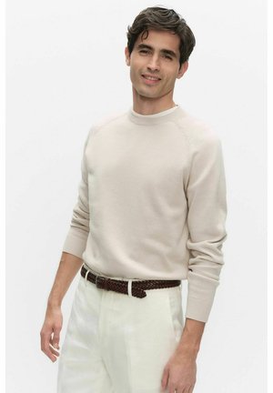 Mężczyzna w beżowym swetrze i białych spodniach z brązowym plecionym paskiem stoi na tle gładkiej białej ściany, delikatnie się uśmiechając.