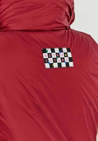 Bordová péřová bunda se čtvercovou nášivkou s černobílým kostkovaným vzorem a vícebarevným textem "RETRO FUTURO".