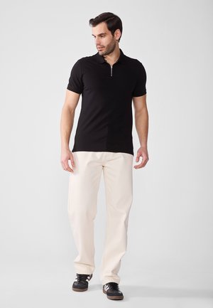 Schwarzes Poloshirt mit kurzen Ärmeln und Reißverschlusskragen, getragen mit cremefarbenen Hosen und schwarzen Sneakers. Glattes Material mit einer taillierten Passform.
