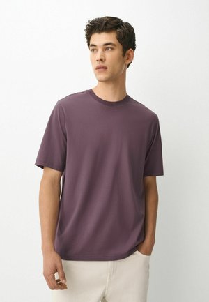 Ung mand med kort krøllet hår iført en ensfarvet lilla kortærmet t-shirt og lyse bukser mod en hvid baggrund.
