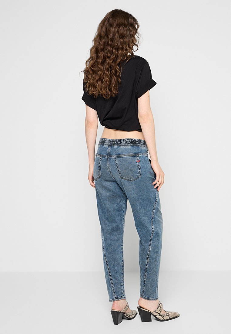 Dunkel geschnittenes T-Shirt, blaue taillierte Jeans mit elastischem Bund, die eine ausgewaschene Textur aufweisen. Das Outfit wird mit gemusterten Blockabsätzen abgerundet.
