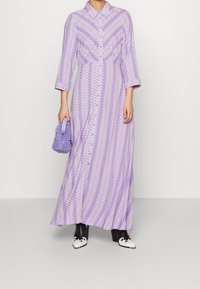 Långärmad lila klänning med vitt zickzackmönster, knappslut framtill och en flödande siluett. Matchad med en fluffig lavendelfärgad väska och svarta stövletter.