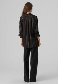 Black button-up shirt with gold vertical stripes, long sleeves rolled at the cuffs, and a relaxed fit. Paired with black wide-leg trousers.