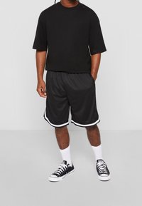 T-shirt noir surdimensionné à manches courtes, associé à un short noir avec des rayures blanches sur les côtés, et des baskets noires avec des semelles blanches.