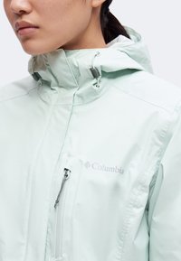Gros plan d'une personne portant une veste Columbia vert clair avec un col montant et une poche poitrine zippée.