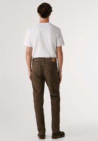 Pantalones de pana marrones con una superficie texturizada, que presentan un parche de piel de la marca en la cinturilla y dos bolsillos traseros, combinados con una camiseta blanca.