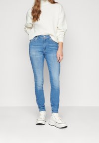 Vaqueros skinny de color azul claro con una cintura de altura media, que presentan un clásico diseño de cinco bolsillos y un leve desgaste, combinados con un suéter crema y zapatillas blancas.