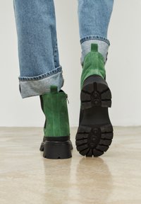 Groene suède enkellaarsjes met een zwarte rubberen zool, uitgerust met een ritssluiting en een gestructureerde zool voor grip. Lichtblauwe denim jeans met een franjeszoom.