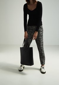 Sac fourre-tout en cuir noir avec deux poignées, design rectangulaire épuré, quincaillerie minimale. Le modèle porte un pantalon à motifs et des chaussures blanches à lacets.