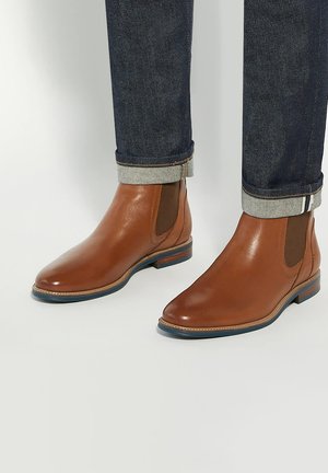 Bottes Chelsea en cuir marron portées avec un jean bleu foncé à revers sur fond blanc.