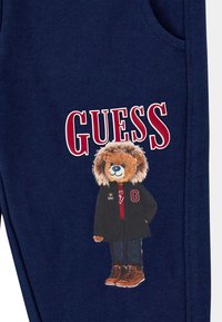 Navyblauwe stof met een cartoonleeuw die een jas en laarzen draagt. Het woord "GUESS" in rode letters is prominent weergegeven boven het ontwerp.