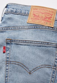 Jean Levi's 515 bleu clair, coupe slim, jambe droite, avec poche arrière ornée d'un patch en cuir camel de la marque et d'une étiquette rouge Levi's sur la couture de la poche.